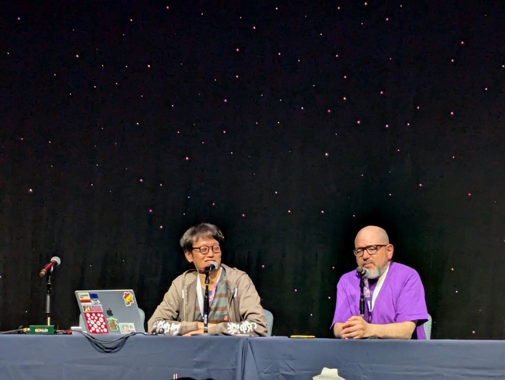 A photo of two men presenting on stage. The person on the left is Kobayashi Hiroyasu. The person on the right is Dan Kanemitsu.