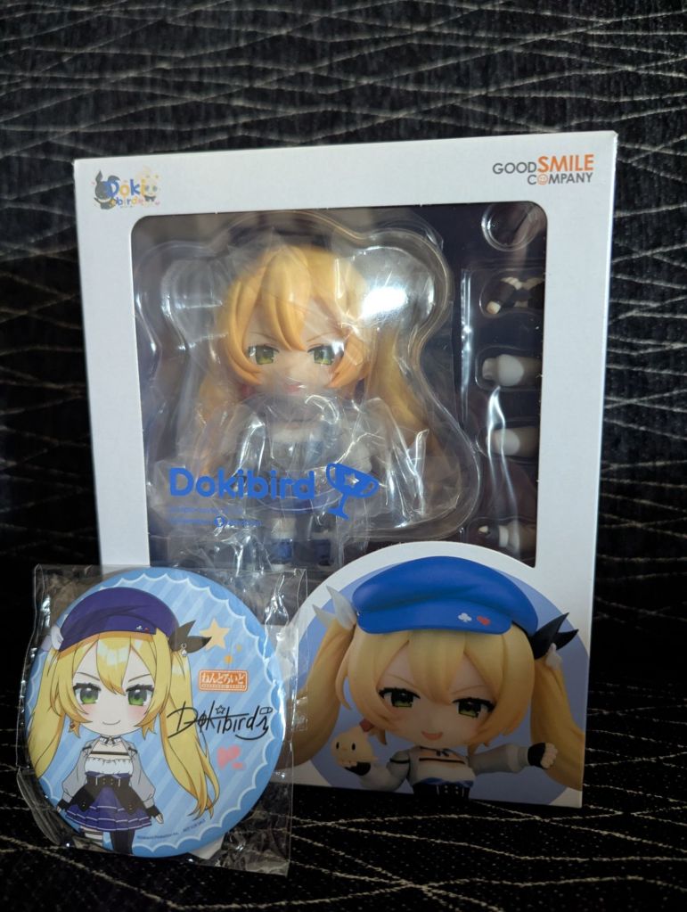 The box for the Nendoroid Dokibird figure, showing a blonde anime girl with long pigtails in a blue dress and beret, a white frilled shirt, and a gray jacket. A metal button featuring Dokibird is in front.