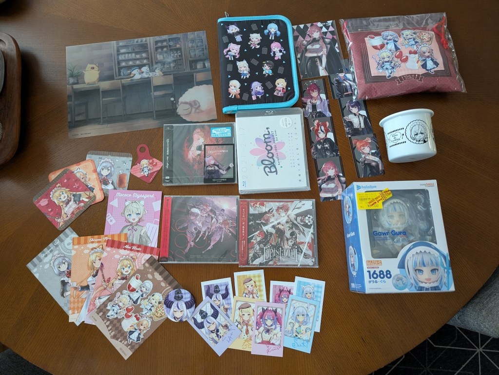 Various hololive merch, including Vesta de Cooking items: a pencil board, a decorative pouch (with chocolate chip cookie inside, hidden), a white metal cup, coasters, and postcard-sized illustrations. Also includes some CDs, a blu-ray, little cards, and a Gawr Gura Nendoroid.