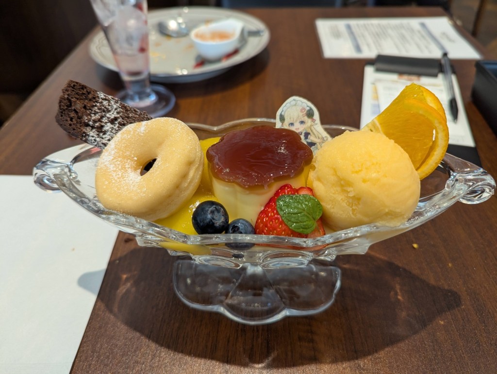 A clear dish with a Japanese-style custard pudding, sorbet, a donut, and various fruits on top, decorated with a little edible Watame wafer.