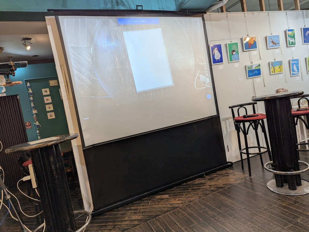 An empty space showing a projection screen and two tall, round tables with tall stools near them.